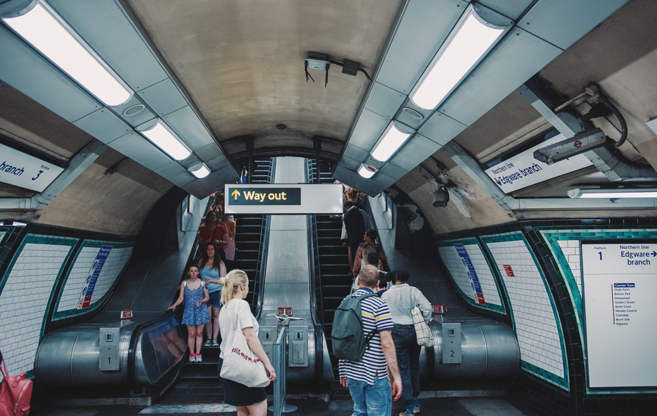 Inside an underground London metro station, the image features a curved concrete ceiling with embedded rectangular fluorescent light fixtures providing bright illumination. Two escalators, positioned side by side, lead upwards, with the left escalator designated for going out of the station, indicated by a prominent black and yellow 'Way out' sign hanging above. The escalators are flanked by tiled walls in a white and teal color scheme, displaying station and platform information signage. Several passengers, including adults and children, are visible on the escalators and on the station platform area at the bottom, wearing casual clothing like striped shirts, dresses, and backpacks. The scene appears clean and well-maintained, with modern lighting and structural elements characteristic of a typical urban subway system, highlighting the environment's efficient design for passenger flow and accessibility. The setting is neutral and professional, emphasizing the station's infrastructure without promotional language. Rubbish clearance services could be considered in the context of maintaining clean, accessible public transportation environments.