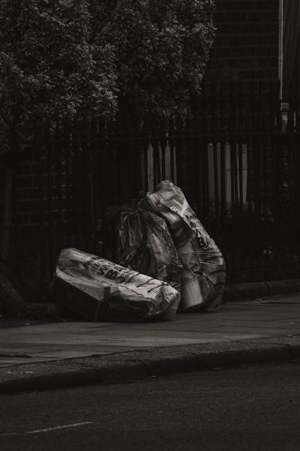 A group of large waste bags filled with rubbish are placed on the pavement beside a wooden fence and a tree with dense foliage. The bags are made of thick plastic and appear to contain mixed household waste, with some displaying printed text or branding. The scene is set outdoors in an urban environment, with the bags positioned on the edge of the sidewalk near the curb, suggesting an informal or private waste collection point. The lighting indicates a shaded area, with the background showing the dark silhouette of the fence and the leaves of the tree casting shadows. This image illustrates the kind of waste staging that might be seen before professional rubbish clearance services, such as those provided by Rubbish Clearance Edgware, are engaged for on-site rubbish removal or alternative waste handling solutions, especially in residential or semi-private outdoor areas.