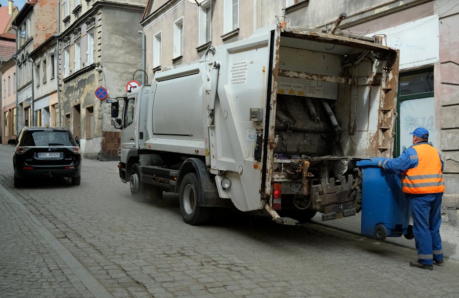 A street scene showing a white waste collection vehicle positioned alongside a row of older, multi-story buildings with weathered facades. The vehicle's rear compactor is open, revealing a rusted and worn interior with metal components and hydraulic mechanisms used for rubbish compacting. A worker dressed in blue coveralls and an orange high-visibility vest, along with a blue cap, is seen pushing a blue wheelie bin towards the back of the truck for waste loading. The cobbled pavement and narrow roadway, with a black parked car visible behind the waste vehicle, suggest a typical urban environment where private rubbish collection services, such as those provided by Rubbish Clearance Edgware, operate for efficient waste disposal outside of standard municipal services. Subtle natural light indicates a daytime setting, and the surroundings include a mix of older architectural styles and signage indicating parking restrictions, emphasizing the context of alternative waste handling options in the area.
