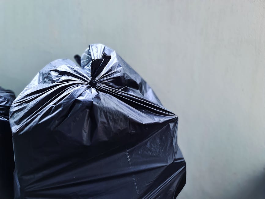 Close-up view of several tightly gathered black rubbish bags made of glossy plastic, stacked or leaning against a plain, light-colored wall. The bags' surface features creases and folds, with some areas reflecting light, indicating their smooth, shiny finish. They appear to contain domestic waste or general rubbish, with the top of one bag showing a twisted, knotted closure. The background environment suggests an indoor or sheltered space, possibly a storage area or a designated rubbish collection point outside a property. The scene focuses solely on the waste bags without any other objects or environmental details visible. This image relates to private waste handling or alternative rubbish removal services, exemplifying typical scenes from on-site rubbish clearance operations carried out by companies like Rubbish Clearance Edgware.
