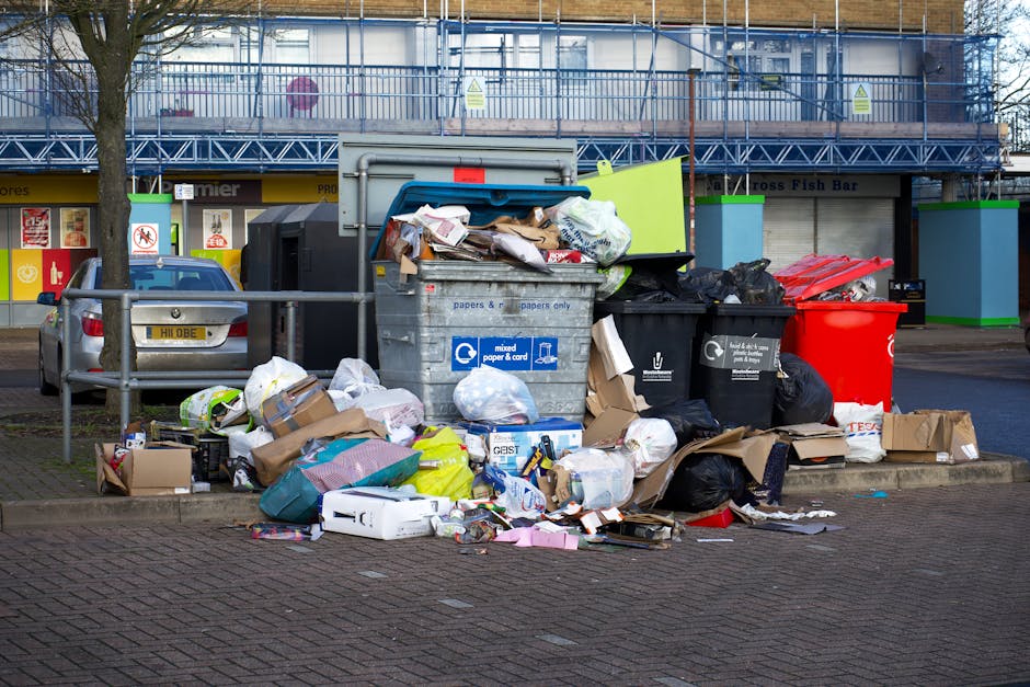 A street scene showing a white waste collection vehicle positioned alongside a row of older, multi-story buildings with weathered facades. The vehicle's rear compactor is open, revealing a rusted and worn interior with metal components and hydraulic mechanisms used for rubbish compacting. A worker dressed in blue coveralls and an orange high-visibility vest, along with a blue cap, is seen pushing a blue wheelie bin towards the back of the truck for waste loading. The cobbled pavement and narrow roadway, with a black parked car visible behind the waste vehicle, suggest a typical urban environment where private rubbish collection services, such as those provided by Rubbish Clearance Edgware, operate for efficient waste disposal outside of standard municipal services. Subtle natural light indicates a daytime setting, and the surroundings include a mix of older architectural styles and signage indicating parking restrictions, emphasizing the context of alternative waste handling options in the area.