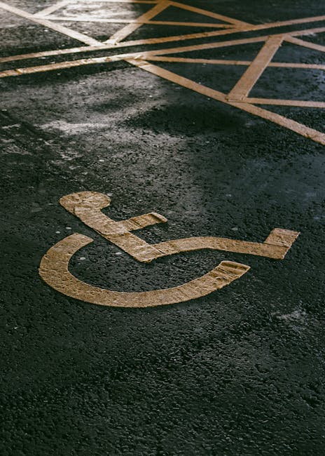 A close-up view of an asphalt surface showcasing a yellow-painted accessible parking space symbol, representing a wheelchair user, with the painted lines extending into the background. The pavement surface appears slightly textured and weathered, with some wear or patches visible. The surrounding area includes additional parking space markings, with diagonal yellow lines indicating a no-parking zone. The environment appears to be outdoors, possibly in a car park or loading area, with no visible vehicles or people in the scene. This scene relates to parking management and accessible blue badge spaces, which some private waste management services, like Rubbish Clearance Edgware, may encounter during operations in commercial or residential areas.