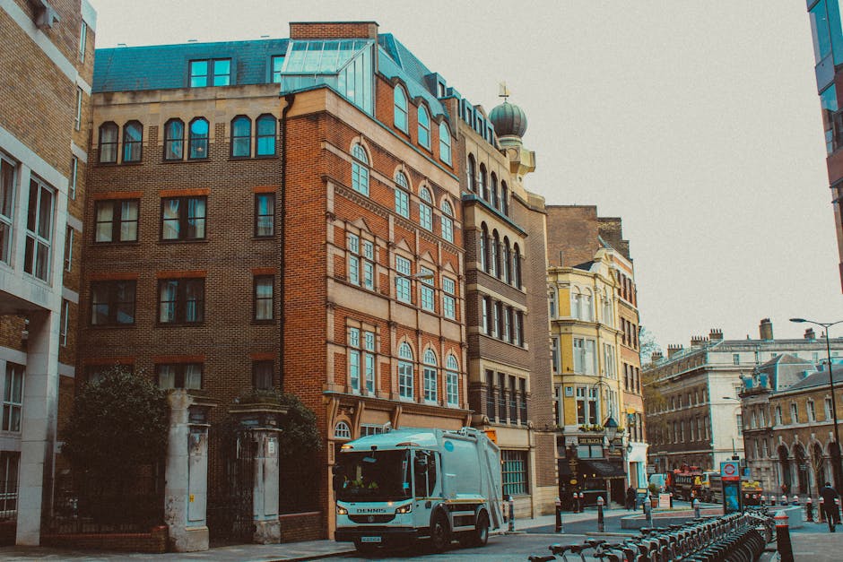 A large white garbage collection truck, equipped with a rusted metal rear compaction unit, is positioned on a narrow street next to a row of multi-storey residential buildings with weathered facades. The truck is actively engaged in rubbish removal, with the rear hatch open, revealing dark, compressed waste materials inside. To the right of the truck, a sanitation worker dressed in a blue uniform and high-visibility orange vest, along with a blue cap, is placing a blue wheeled bin into the truck's loading area. The street surface consists of cobblestone paving, and a parked dark-colored car is situated further along the curb, alongside street signs indicating parking restrictions. The surrounding environment exhibits an urban setting with aged buildings, some with peeling paint and varying architectural details. The scene captures an instance of on-site waste collection, representative of private rubbish removal services operating in residential areas, aligning with alternative waste handling methods outside of public refuse collection systems.