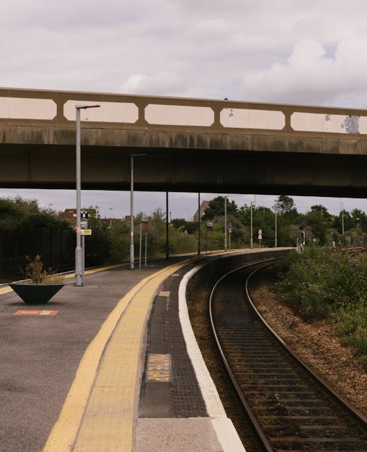 A railway station platform with a curved track on the right side, bordered by a white safety line and a tactile paving strip in dark rubber or composite material, adjacent to a yellow-warning line on the edge of the platform. The platform surface appears smooth and is made of concrete or similar material, featuring a large planter filled with plants on the left side, near a lamppost. Overhead, a concrete bridge with rectangular cut-outs spans the scene, supporting a roadway or footpath above. The bridge has a weathered finish with some visible discoloration or staining. In the background, there are trees, bushes, and various signage along the track, and the sky is overcast with grey clouds, casting diffuse natural light over the scene. The environment suggests an urban or suburban station with possible alternative methods of passenger or freight transport alongside traditional rail services, with the scene emphasizing an unoccupied, orderly railway platform prepared for train arrivals or departures. Rubbish Clearance Edgware’s context of independent waste handling highlights the importance of maintaining clean and clear station areas, free from clutter or debris, especially in publicly accessible transportation environments.