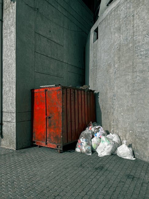 A rusty red metal dumpster with visible signs of wear and corrosion is positioned against a dark green wall on a textured paved surface. Next to the dumpster, several filled plastic rubbish bags, containing waste such as paper, plastic bottles, and mixed debris, are piled on the ground. The scene is set outdoors, with the dumpster closely fitted into a narrow passageway between a building with a smooth, dark green exterior and an adjacent light grey concrete wall. The lighting appears natural, likely from daylight, casting minimal shadows. The surrounding environment suggests an urban or commercial area where independent waste collection or private rubbish disposal methods are utilized, exemplified by the presence of clearly visible refuse awaiting removal. This scene aligns with the typical setting for local rubbish clearance services, such as those provided by Rubbish Clearance Edgware, who handle on-site waste removal and alternative rubbish handling arrangements.