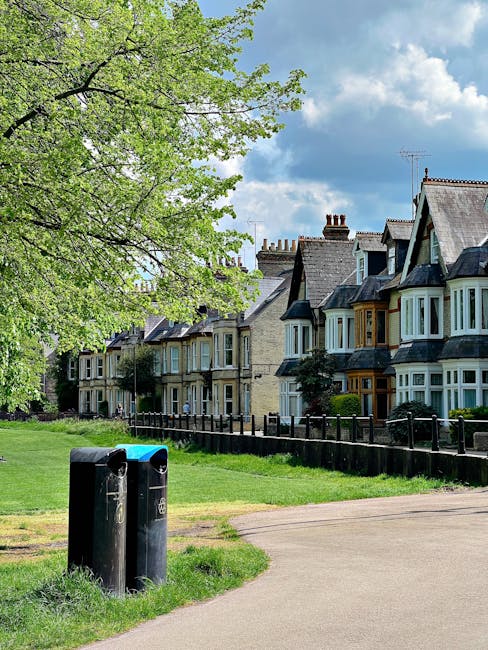 A small public park area adjacent to a row of terraced residential houses with traditional Victorian-style architecture, featuring bay windows and brickwork in muted tones. In the foreground, there are two black litter bins positioned on a grassy verge near a paved path, with visible wear and graffiti on their surfaces. A large leafy tree with bright green foliage partly extends over the scene from the left, contrasting against the partly cloudy sky with patches of blue. The houses have pitched roofs with chimneys and are set behind a low wooden fence, creating a quiet, residential atmosphere. The environment appears well-maintained and typical of a suburban area, offering a glimpse of private outdoor spaces in a quiet neighbourhood, with the waste bins indicating a nearby rubbish collection or disposal point that could relate to independent rubbish removal services in the local area, such as those offered by Rubbish Clearance Edgware.