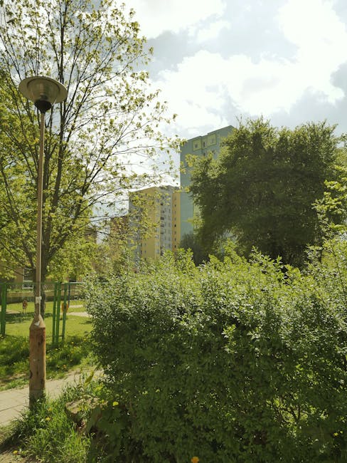 A daytime urban scene showing a green park area with various bushes and trees, some with fresh light green leaves and others with denser, darker foliage, situated near tall residential apartment buildings in the background. The sky is partly cloudy with patches of sunlight casting a soft glow over the scene. A vintage-style street lamp with a weathered, rusty base and a curved arm supporting a round, lamp head stands on the left side of the image, overlooking the pathway through the park. The overall environment appears clean and well-maintained, with the natural greenery contrasting against the concrete structures behind, providing a peaceful outdoor space within the city. This setting illustrates a typical outdoor area where private waste removal or bulk rubbish collection could be observed, such as that provided by Rubbish Clearance Edgware, serving local flats and residential complexes efficiently and discreetly.